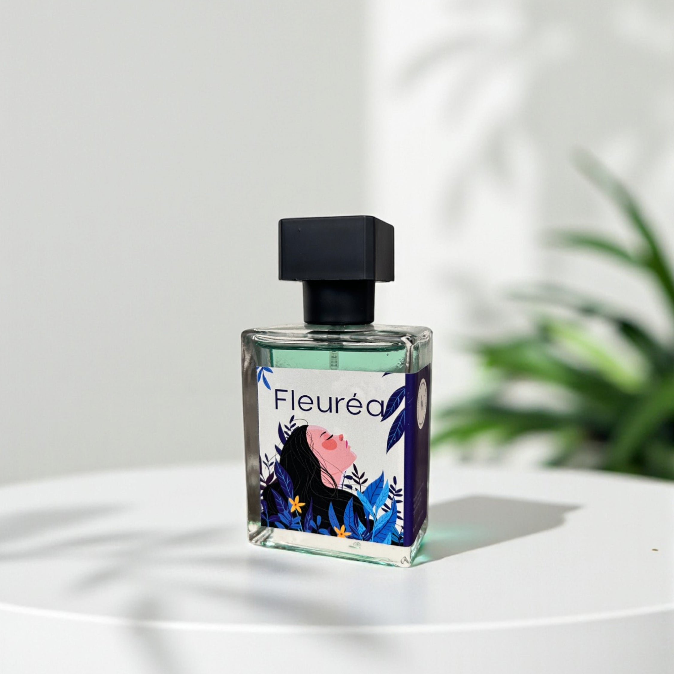 "Fleureá perfume, square glass bottle with black cap on a white table, soft plant in the background"